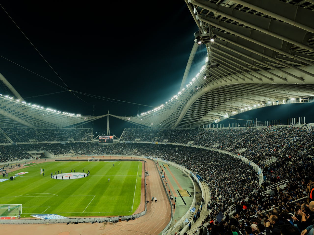 Een vol voetbalstadion onder felle verlichting in de avond, waarop de spanning van een live sportevenement wordt vastgelegd.